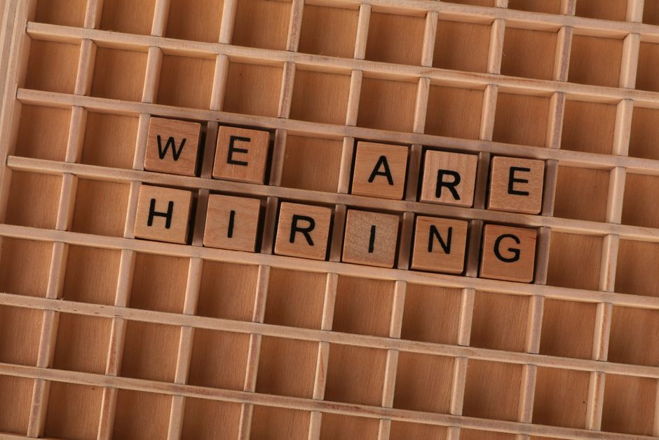 A close-up view of a wooden letter board with small square compartments, placed on a light wooden surface. The board displays the message 'WE ARE HIRING' arranged with individual black letter tiles. T